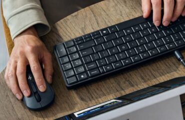 Primer plano de una persona utilizando un teclado HP negro ultradelgado y un ratón inalámbrico HP sobre un escritorio de madera. El teclado está conectado mediante un cable USB-C a un monitor parcialmente visible, mostrando un entorno de trabajo moderno y minimalista