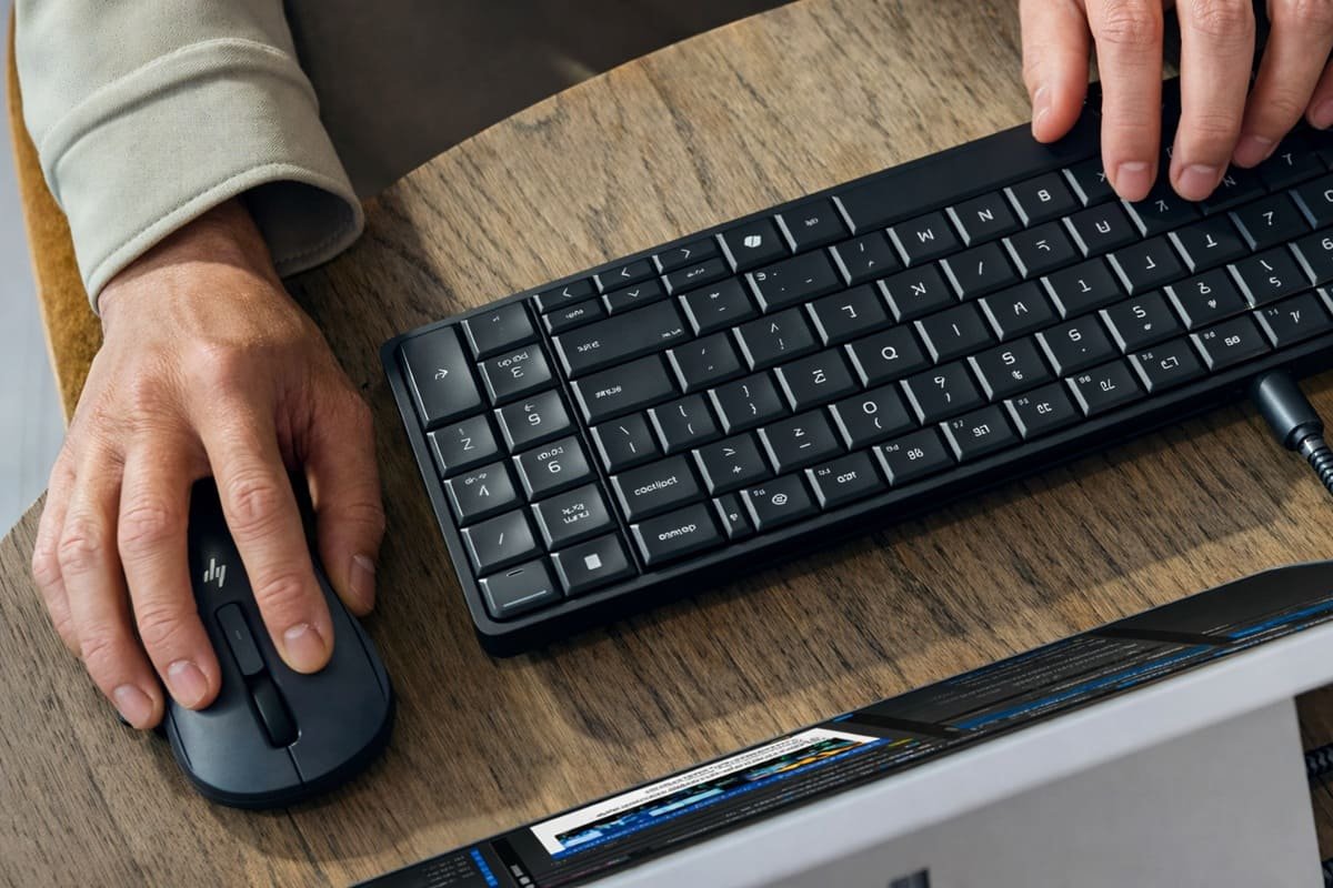 Primer plano de una persona utilizando un teclado HP negro ultradelgado y un ratón inalámbrico HP sobre un escritorio de madera. El teclado está conectado mediante un cable USB-C a un monitor parcialmente visible, mostrando un entorno de trabajo moderno y minimalista