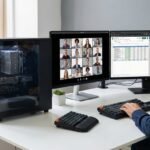 etup de teletrabajo con un PC de torre sobrio y un disipador de aire visible, junto a dos monitores que muestran una videollamada y hojas de cálculo, representando el equipo ideal para productividad.
