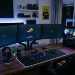 Una persona sentada frente a un moderno setup de PC en una habitación con iluminación ambiental azul y morada. El amplio escritorio de madera cuenta con un sistema de tres monitores que muestra la pantalla de carga del sistema, un teclado y ratón con luces RGB sobre una gran alfombrilla iluminada, y una torre de ordenador con panel de cristal que deja ver los componentes y ventiladores brillantes en su interior. Al fondo se aprecian estantes decorados con figuras coleccionables y paneles acústicos