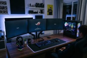 Una persona sentada frente a un moderno setup de PC en una habitación con iluminación ambiental azul y morada. El amplio escritorio de madera cuenta con un sistema de tres monitores que muestra la pantalla de carga del sistema, un teclado y ratón con luces RGB sobre una gran alfombrilla iluminada, y una torre de ordenador con panel de cristal que deja ver los componentes y ventiladores brillantes en su interior. Al fondo se aprecian estantes decorados con figuras coleccionables y paneles acústicos