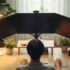 Persona sentada frente a una mesa de trabajo con tres monitores, una lámpara y plantas decorativas.
