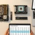 Conjunto de componentes de computadora, incluyendo una placa base, un procesador, una memoria RAM y un laptop, mientras se consulta información de compatibilidad en una tablet.