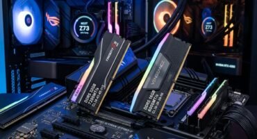 Dos sticks the memoria RAM expuestos encima de una placa base gaming con iluminacion rgb. Y dos PCs gaming de fondo.