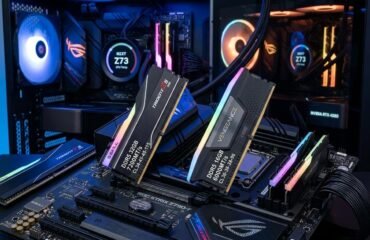 Dos sticks the memoria RAM expuestos encima de una placa base gaming con iluminacion rgb. Y dos PCs gaming de fondo.