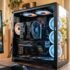 Torre de PC de escritorio con panel de vidrio templado que muestra componentes internos, un gran disipador de CPU y varios ventiladores con iluminación RGB en tonos azules y naranjas. El equipo está sobre un escritorio de madera junto a una ventana iluminada por la luz natural del verano.