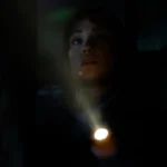 Una joven ilumina la oscuridad con una linterna mientras mira hacia arriba con gesto de alerta.