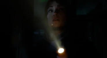 Una joven ilumina la oscuridad con una linterna mientras mira hacia arriba con gesto de alerta.