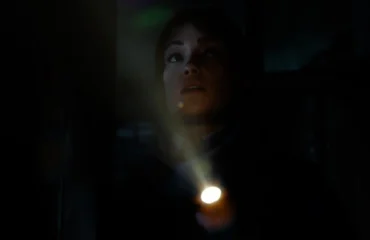 Una joven ilumina la oscuridad con una linterna mientras mira hacia arriba con gesto de alerta.