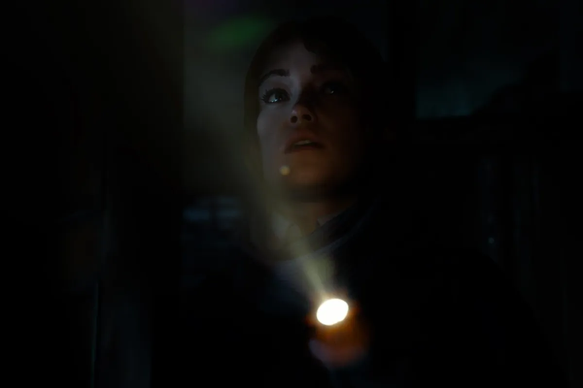 175-until-dawn-2-ps5-fecha-lanzamiento Una joven ilumina la oscuridad con una linterna mientras mira hacia arriba con gesto de alerta.