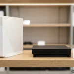 Dos consolas de sobremesa, una blanca vertical y otra negra, expuestas en la estantería de una tienda. Imagen generada con IA.