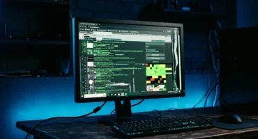Monitor mostrando código fuente y gráficos de píxeles de colores en una mesa con teclado retroiluminado. Imagen generada con IA.