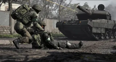 Captura oficial de Wardogs: un soldado arrastra a un compañero herido por una calle embarrada mientras un tanque avanza detrás.