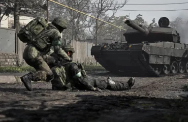 383-wardogs-millon-copias-300000-jugadores-steam Captura oficial de Wardogs: un soldado arrastra a un compañero herido por una calle embarrada mientras un tanque avanza detrás.