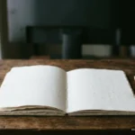 Cuaderno abierto en blanco sobre una mesa de madera, con un lápiz al lado y una pantalla apagada al fondo. Imagen generada con IA.