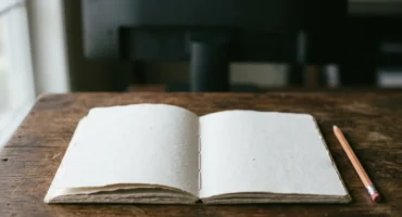Cuaderno abierto en blanco sobre una mesa de madera, con un lápiz al lado y una pantalla apagada al fondo. Imagen generada con IA.