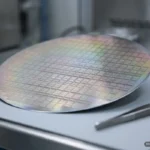 Una oblea de silicio redonda y sin marcas, inclinada sobre una mesa clara de laboratorio, con reflejos irisados de arcoíris sobre su cuadrícula de pequeños rectángulos idénticos; a la derecha, unas pinzas metálicas apoyadas y, al fondo, un laboratorio desenfocado. Imagen generada con IA.