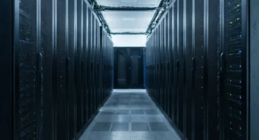 Pasillo de un centro de datos entre dos filas de armarios de servidores negros con pequeñas luces de estado verdes y azules. Imagen generada con IA.