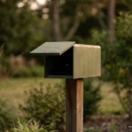 Un buzón metálico verde oliva, liso y sin marcas, sobre un poste de madera en un jardín al atardecer, con la tapa entreabierta y un diminuto punto de luz azul en su interior oscuro; setos, hierba y árboles desenfocados al fondo. Imagen generada con IA.