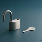 Un candado de acero mate abierto y sin marcas sobre una superficie lisa de color azul petróleo, con una llave plateada sencilla al lado y una luz lateral suave que proyecta sombras largas. Imagen generada con IA.