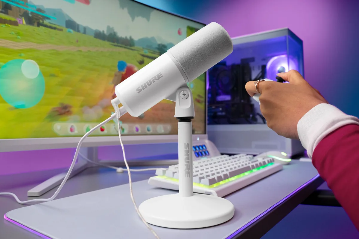shure-mv6-gen-2-blanco El micrófono Shure MV6 Gen 2 en blanco, sobre su soporte de escritorio, junto a un teclado, un monitor con un juego y un PC con iluminación morada