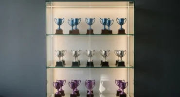 Vitrina de cristal iluminada con tres baldas de trofeos de copa, azules arriba, plateados en medio y morados abajo, con un hueco libre. Imagen generada con IA.