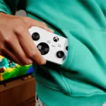 Xbox Nano Controller: una mano guarda el mando, blanco y compacto, en el bolsillo de una sudadera verde, delante de un televisor con un videojuego en pantalla