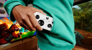 Xbox Nano Controller: una mano guarda el mando, blanco y compacto, en el bolsillo de una sudadera verde, delante de un televisor con un videojuego en pantalla