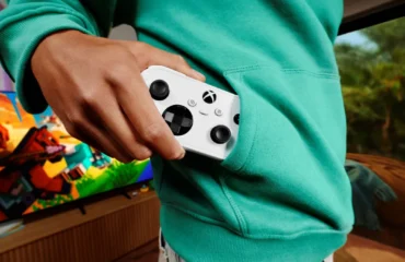 xbox-nano-controller Xbox Nano Controller: una mano guarda el mando, blanco y compacto, en el bolsillo de una sudadera verde, delante de un televisor con un videojuego en pantalla